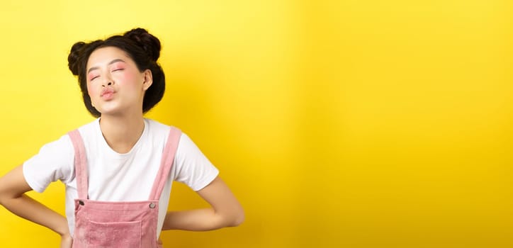 Romantic korean girl close eyes and pucker lips, waiting for sweet kiss on Valentines day, kissing lover, standing on yellow background.