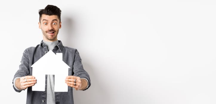 Real estate and insurance concept. Man look with excitement at paper house cutout, buying home, standing on white background.