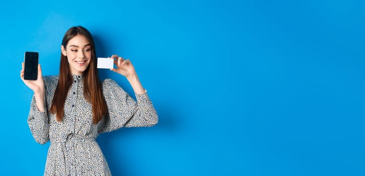 Online shopping. Beautiful female model showing empty cellphone screen and plastic credit card, smiling pleased, buying in internet store, blue background.