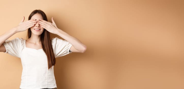 Happy romantic girl covering eyes and waiting for surprise, smiling while anticipating gift, standing on beige background.