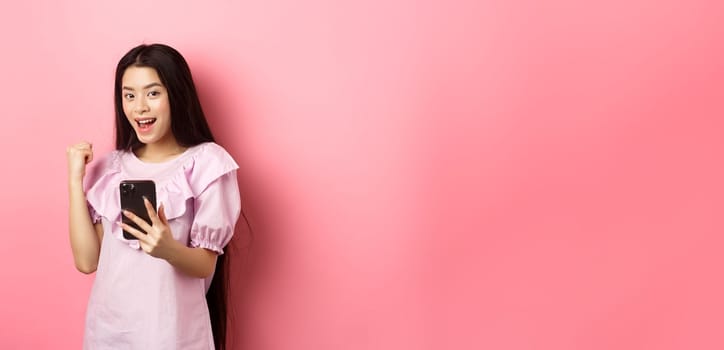 Online shopping. Satisfied asian girl winning on mobile phone, say yes and make fist pump, holding smartphone, standing against pink background.