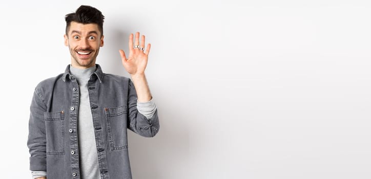 Friendly stylish man saying hello and waiving hand, smiling cheerful, greeting you with hi gesture, standing on white background.