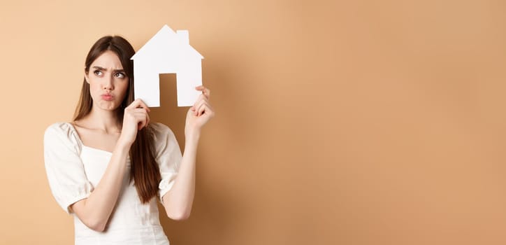 Real estate. Upset young woman showing paper house cutout and frowning sad, looking aside thoughtful, thinking of buying apartment, standing on beige background.