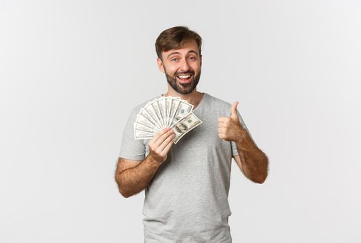 Portrait of handsome man with beard, showing thumbs-up and holding money, recommend credit or loan, standing over white background.