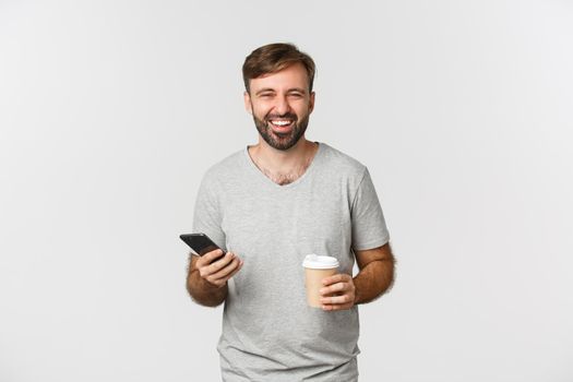 Portrait of carefree attractive man, drinking coffee and using mobile phone, laughing and smiling, standing over white background.