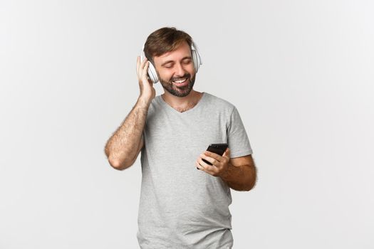 Image of handsome man in casual outfit, listening music in wireless headphones, holding mobile phone, smiling pleased, standing over white background.