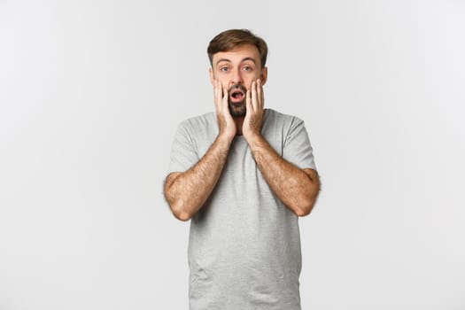Image of bearded man in casual grey t-shirt, gasping amazed, looking at something impressive, standing over white background.