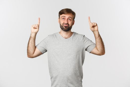 Skeptical and unimpressed adult man with beard, looking up and pouting displeased, standing over white background in gray t-shirt.