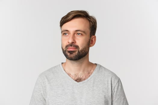 Portrait of handsome bearded man in gray t-shirt, looking left thoughtful, standing over white background.