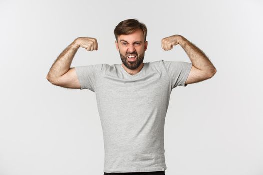 Portrait of confident bearded man flexing biceps, showing his muscles after gym workout, standing over white background.