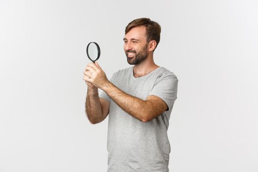 Image of handsome caucasian man in gray t-shirt, searching for something, looking pleased through magnifying glass with curious face, standing over white background.