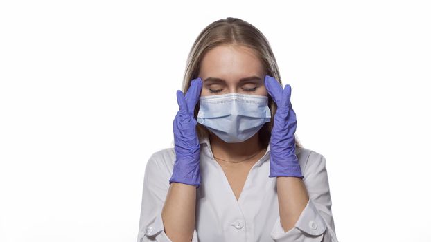 Suffering headache nurse in a medical mask and disposable gloves at work, massaging the temporal lobes of the head, with eyes shut. Isolated on white background. Medicine concept.