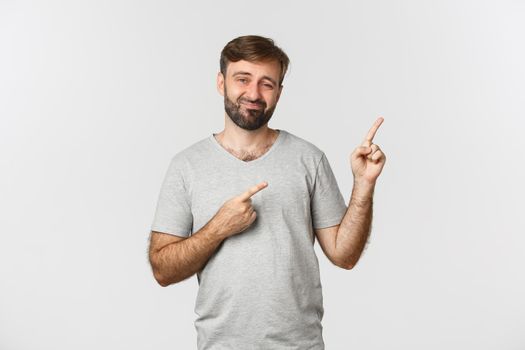 Image of unsure bearded guy in grey t-shirt, having doubts and pointing fingers at upper right corner, standing over white background.