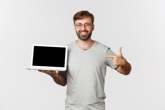 Cheerful bearded man in gray t-shirt, showing laptop screen and smiling, working from home, making thumbs-up in approval, standing over white background.