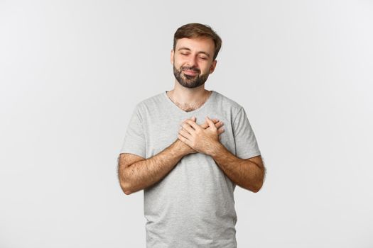 Image of touched bearded man in basic t-shirt, close eyes and holding hands on heart, feeling nostalgic, thinking about something and smiling, standing over white background.