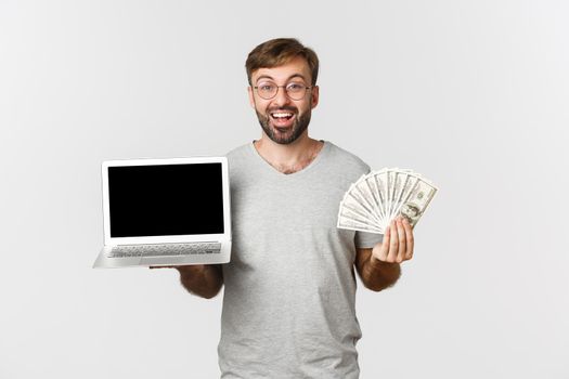 Portrait of excited caucasian man in gray t-shirt, holding laptop and money, showing something on screen, standing over white background.