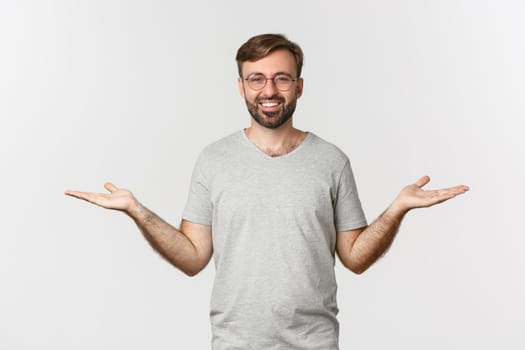 Handsome man with beard, wearing gray t-shirt and glasses, spread hands sideways and holding two things, demonstrate products, standing over white background.