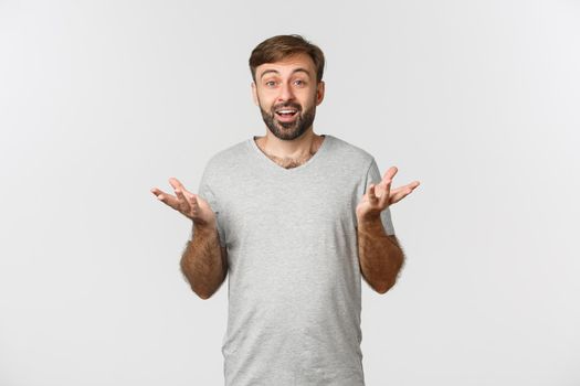 Portrait of handsome bearded male moden in grey t-shirt, raising hands up and looking amazed, congratulating you, standing over white background.