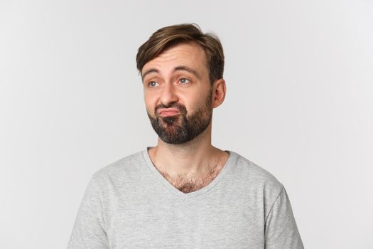Close-up of skeptical caucasian guy with beard, looking upset left and pouting, standing over white background.