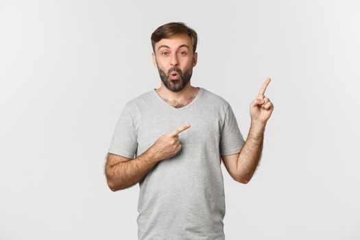 Portrait of handsome surprised guy with beard, wearing gray t-shirt, saying wow and pointing fingers at upper right corner, standing over white background.