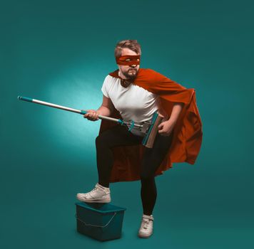 Superhero rock musician is cleaning. Man wearing a red cloak and mask holds a mop representing himself a musician putting his foot on a bucket. Cut out on biscay green background.