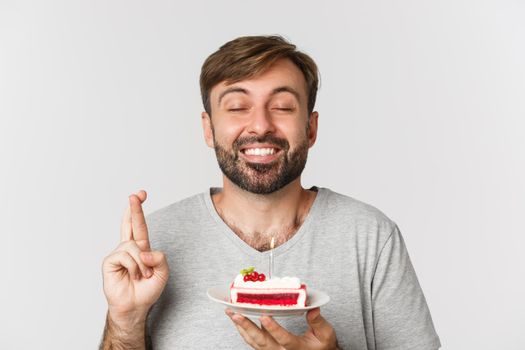 Close-up of hopeful man celebrating birthday, making wish with fingers crossed, holding bday cake, standing over white background.