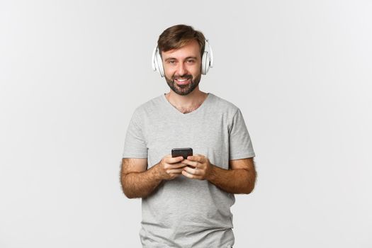 Adult caucasian guy smiling, listening music in headphones and using mobile phone, standing over white background.