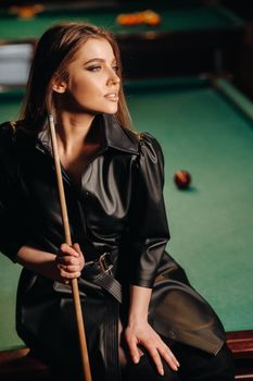 A girl with a cue in her hands sits on a table in a billiard club.Russian billiards.