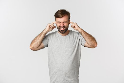 Portrait of annoyed guy in gray t-shirt, shutting ears and grimacing from loud noise, standing over white background.