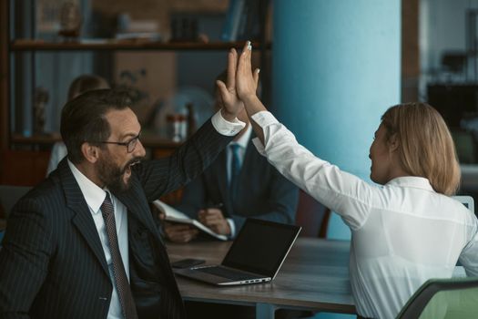 Office workers give high five each other. Happy business man and woman join their hands together sitting at office table with laptop computer on it. Toned image.