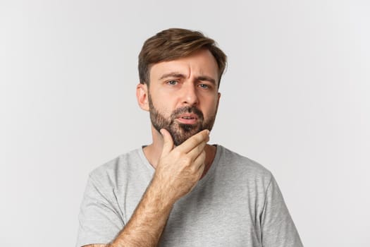 Close-up of confused man touching his beard and looking at himself, need to shave, standing over white background.