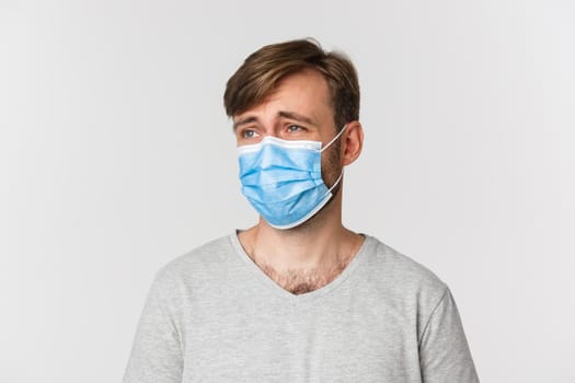 Concept of pandemic, covid-19 and social-distancing. Close-up of handsome man in medical mask, looking left at logo with skeptical and disappointed face, standing over white background.