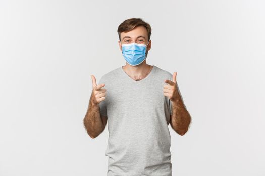 Concept of pandemic, coronavirus and social-distancing. Excited handsome man in gray t-shirt and medical mask, showing thumbs-up and smiling, liking something good.