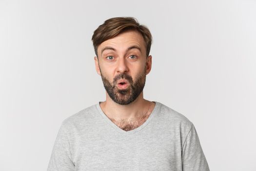 Close-up of handsome adult man with beard, wearing gray t-shirt, looking surprised and curious at advertisment, standing over white background.
