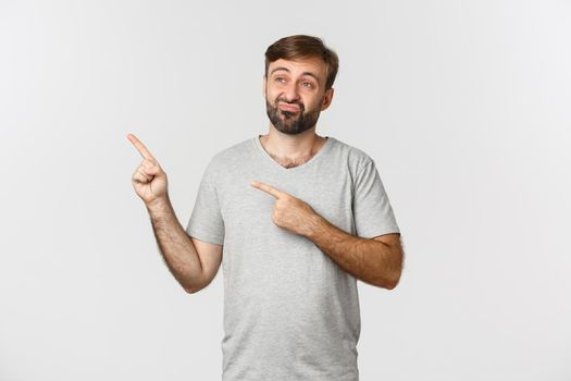 Image of skeptical and unamused bearded guy in gray t-shirt, pouting and pointing fingers at upper left corner, standing over white background.