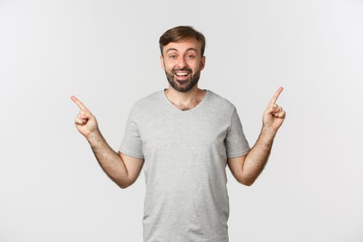 Portrait of happy smiling man with beard in gray t-shirt, pointing fingers sideways, showing two good choices, standing over white background.