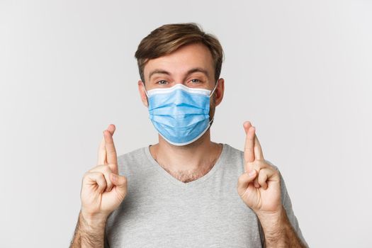 Concept of pandemic, covid-19 and social-distancing. Hopeful worried guy in medical mask, cross fingers for good luck and making wish, standing over white background.