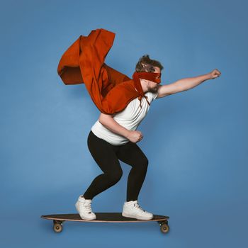 Super speed concept. Superhero rushes forward on a skateboard. Man in red superhero costume with a flying cloak isolated on faded denim blue background.