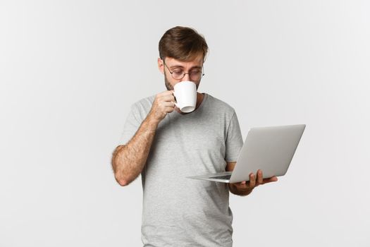 Image of male freelancer working with laptop, drinking coffee, standing in gray t-shirt and glasses over white background.