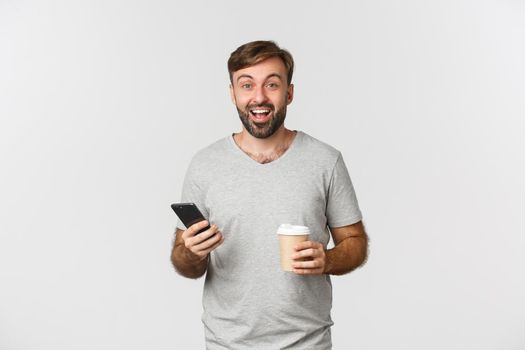 Image of modern caucasian man with beard, drinking coffee and casually using mobile phone, standing excited over white background.