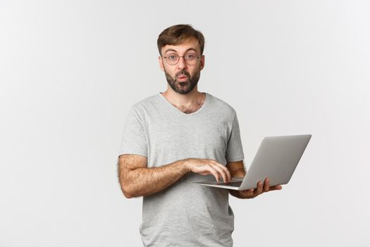 Image of surprised male model in glasses and gray t-shirt, looking amazed, using laptop, standing over white background.