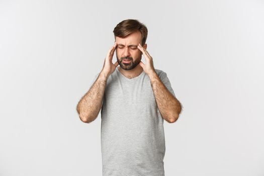 Man having a severe headache, grimacing and touching head, feeling dizzy or sick, standing in gray t-shirt over white background.