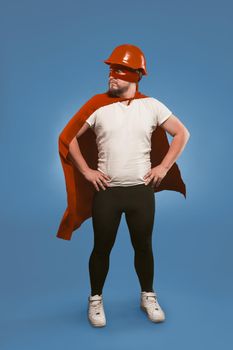 Super hero engineer. Man wearing red helmet, mask and cape standing with his hands on hips. Isolated on faded denim blue background.