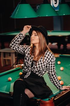 A girl in a hat in a billiard club sits on a billiard table.Playing pool.