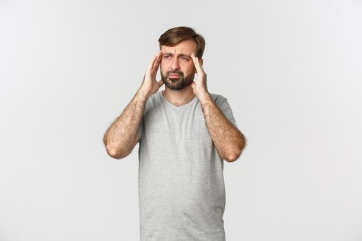 Man having a severe headache, grimacing and touching head, feeling dizzy or sick, standing in gray t-shirt over white background.