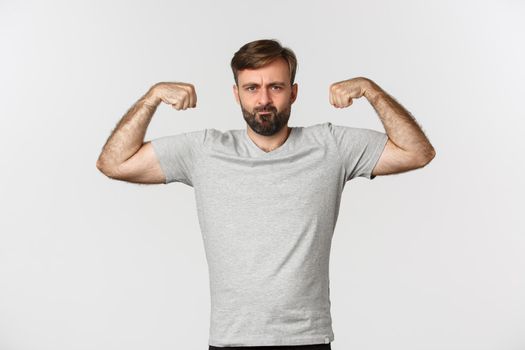 Portrait of confident bearded man flexing biceps, showing his muscles after workout, standing over white background.