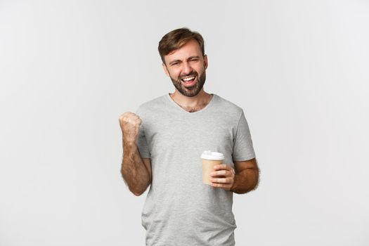Portrait of handsome man with beard, wearing gray basic t-shirt, feeling energized after drinking coffee, making fist pump and saying yes with excitement, standing over white background.