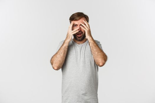 Portrait of embarrassed bearded man in gray t-shirt, cover eyes with hands but peeking through fingers scared to look, standing over white background.