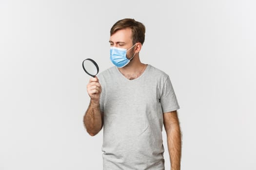 Concept of pandemic, covid-19 and social-distancing. Image of serious man in medical mask, searching for something and looking through magnifying glass, standing over white background.