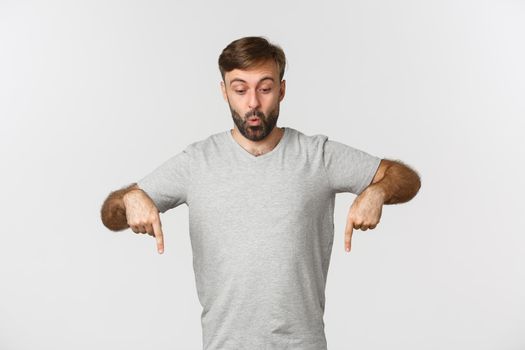 Surprised attractive guy in gray t-shirt, pointing and looking down fascinated, saying wow, standing over white background.